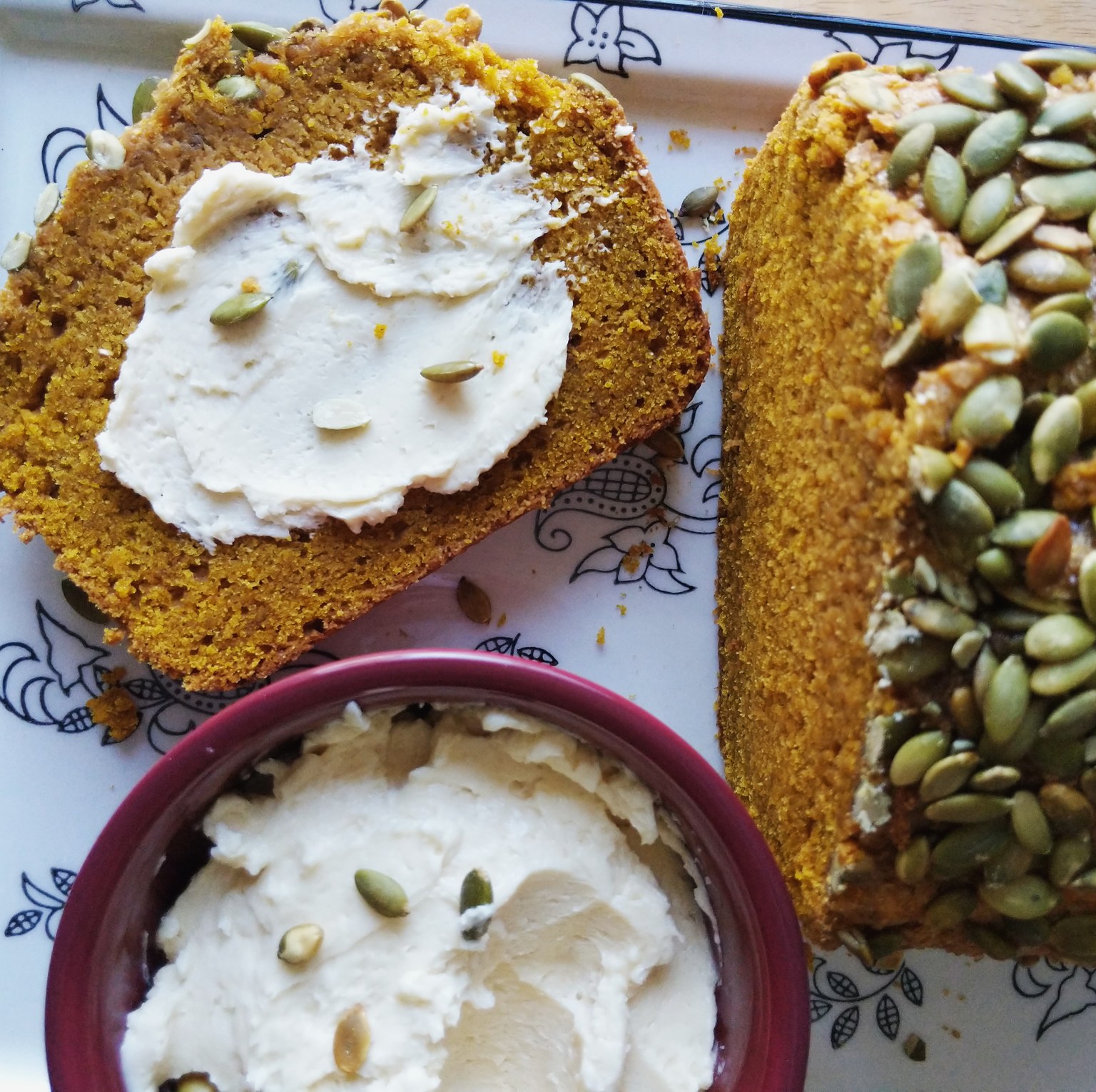 Slices of pumpkin bread with a crunchy pepita crust, one spread with whipped butter, beside a bowl of whipped butter